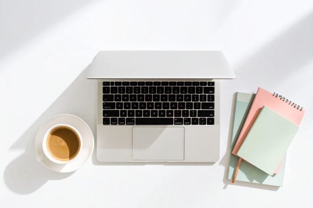 Slighly open MacBook on a white desk with a cup of coffee and stack of notebooks
